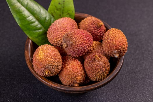 Ripe lychees and green leaves presented in a wooden bowl, creating a vibrant display against a dark backdrop