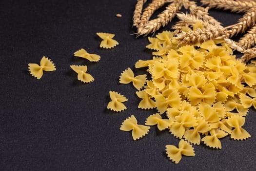 Dry farfalle pasta spilling onto a dark surface, accompanied by wheat ears, evokes a sense of rustic culinary preparation