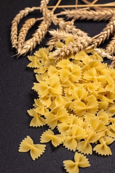 Dry farfalle pasta is scattered on a black surface, accompanied by wheat ears, highlighting the connection between raw ingredients and the final product