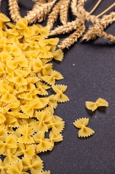 Raw farfalle pasta spilling onto a dark surface, accompanied by wheat ears, evokes the essence of italian cuisine and the journey from grain to gourmet delight