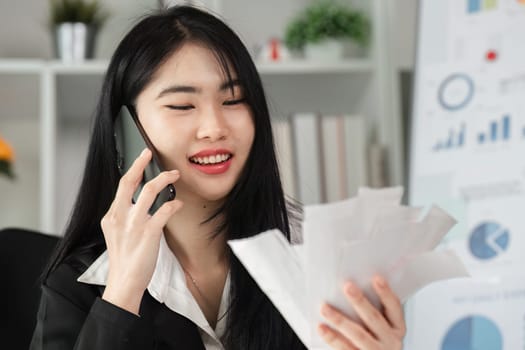 A cheerful businesswoman on a phone call reviews financial documents, showcasing effective communication and multitasking.
