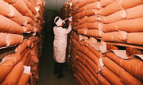 A worker in a white lab coat carefully labels and organizes brown paper-wrapped sugar bags in a well-lit warehouse during daylight hours.
