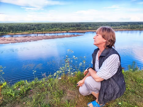 A middle-aged blonde woman kneels at a scenic riverside, capturing a selfie during her travel excursion in nature.