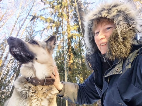 A blonde woman captures a joyful winter selfie with her German Shepherd while exploring a snowy forest.