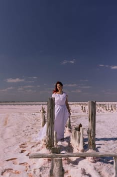 A woman in a white dress walks across a wooden structure on a salt flat. The sky is blue and there are clouds.