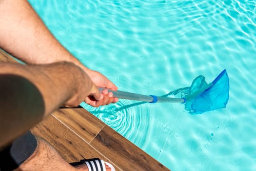 Closeup of man cleaning turquoise swimming pool water with skimmer net and wooden deck