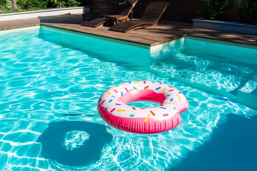 Pink donut-shaped inflatable float in turquoise swimming pool with wooden deck and sunny reflections