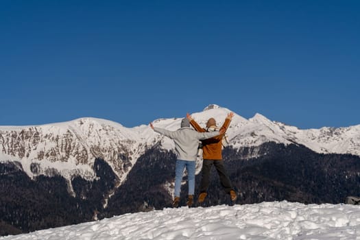 Couple joyfully leaps in snowy mountain landscape. Arms raised, bright clothing contrasts with white snow and dark, snow-capped peaks under a clear blue sky. Winter wonderland fun.