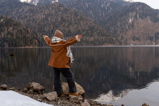 Woman in brown coat stands on rocks by a calm lake, arms outstretched, facing a snow-dusted mountain range. Winter landscape.