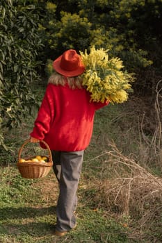 Woman in red sweater and hat carries a large bouquet of yellow mimosa and a basket of fruit, walking away from the camera through a wooded area.
