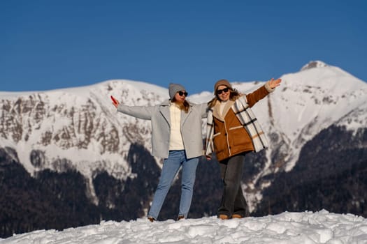 Two women joyfully pose on a snow-covered mountain peak, arms outstretched, against a backdrop of majestic snow-capped mountains under a clear blue sky.