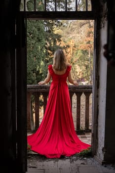 Woman in a long red dress stands on a dilapidated balcony overlooking autumnal foliage. The view is framed by an aged doorway.
