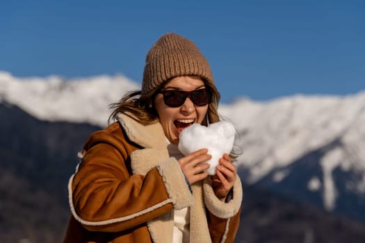Woman in brown coat and beanie playfully bites a heart-shaped snowball. Snowy mountains form a backdrop on a sunny day.