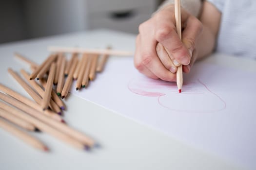 Child drawing heart with colored pencils on white paper.