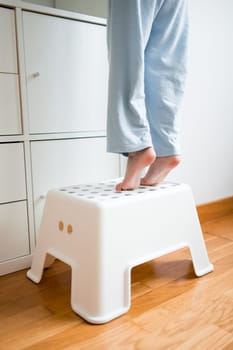 Child standing on white step stool reaching high shelf in home setting.