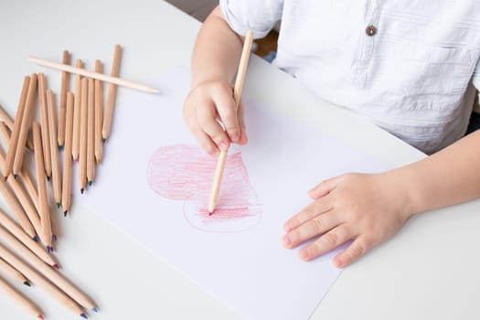 Young caucasian child drawing heart with colored pencils on paper.