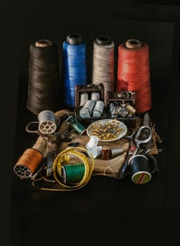 Thimbles in small wooden treasure chest, Spools of thread of various colour, Sewing needles stuck in a green miniature spools thread, Steel scissors and Measuring tape including pins in small round tray on Sackcloth with Black background. Sewing items set or Sewing notions (Sewing objects), use it as your Wallpaper, Poster and Copy space, Selective focus.