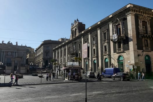 Catania. Italy - March 3, 2025: Iconic Palazzo Tezzano with intricate architecture in Catania's bustling plaza. Features locals, a quaint coffee kiosk, and parked cars under a vivid blue sky.