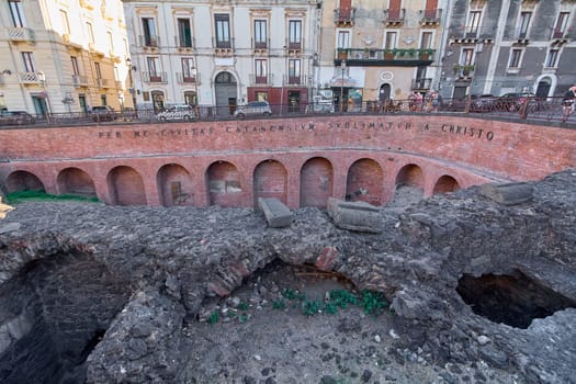 Catania. Italy - March 3, 2025: Stunning Roman Amphitheater excavation with ancient arches and text, set against Catania's vibrant cityscape. Perfect for cultural exploration, historical studies.