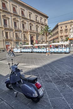 Catania. Italy - March 3, 2025: Lively Catania street with a colorful tour train and parked scooter, surrounded by historic buildings. Ideal for urban travel tourism, and street life photography under