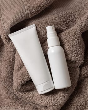 White blank squeeze cream tube and spray bottle on brown towel in bathroom top view, brand packaging mockup, copy space. Natural handmade cosmetic product for facecare and spa procedures