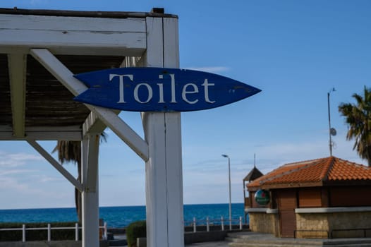 A rustic blue sign indicates restrooms near a vibrant seaside location on a clear day.