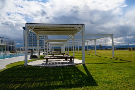 A spacious green area has shaded benches, with mountains and a pool in the distance beneath a cloudy sky.