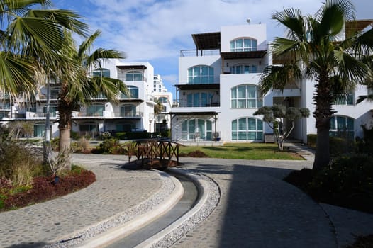 Bright blue skies welcome pedestrians amidst sleek white buildings and lush greenery in a tranquil coastal area.