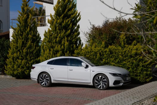 Gaziveren Cyprus -01.03.2025 A sleek white sedan is parked beside tall evergreens, basking in the warmth of a sunny afternoon sky.