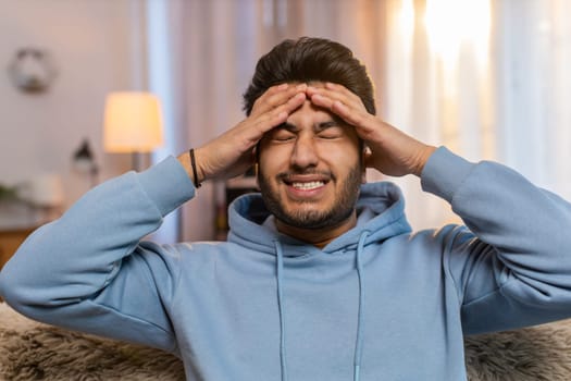 Sick Indian man rubbing temples to cure headache, suffering from tension, migraine, stress, and high blood pressure at home. Hispanic guy holds head, grimacing in pain, experiencing severe discomfort.