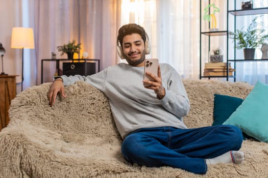 Young Indian man on sofa wearing wireless headphones listening to music on smartphone selects favorite track relaxes and dances. Arabian guy enjoys rhythm moving with energy and cheerful expression.