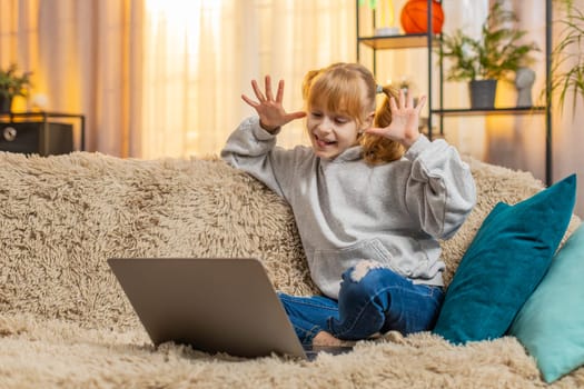 Young teenager girl rests at home with laptop, making a fun video call online and laughing at her own reflections making silly faces. Child kid waves hi in excited joy and saying I am so funny look