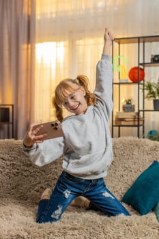 Young teenager girl using smartphone at home, giggling while playing passing a tough online game level with excitement. Child kid on sofa waving phone in joy, celebrate win victory cheerful triumph