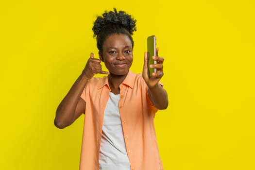 African American woman holds smartphone and makes call me gesture, signaling a desire for connection. Black girl on yellow background radiates confidence and highlights direct invitation to contact