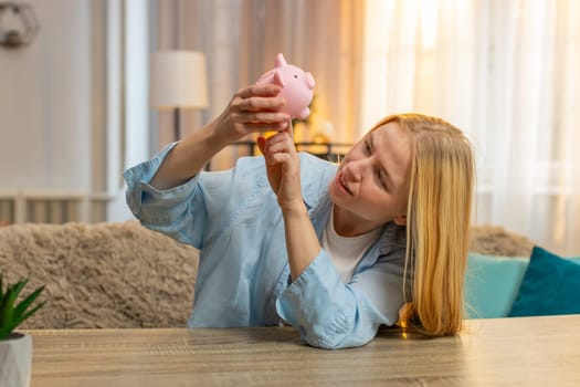 Frustrated young woman trying to get money cash at home, knocking on piggy bank with worried upset face. Caucasian girl sitting at table realizing bankruptcy, showing financial lack. Sunset light