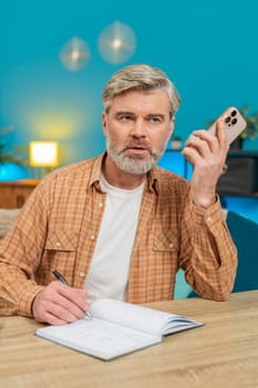 Mature man talking on mobile phone loudspeaker, making business discussion and writing notes in notebook. Middle-aged guy sitting at home table focusing on solutions, feeling determined. Vertical