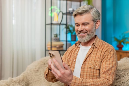 Caucasian mature man sitting on home sofa using smartphone to browse photo social media gallery. Middle-aged guy smiles from pleasant memories and presses like on favorite pictures enjoying the moment