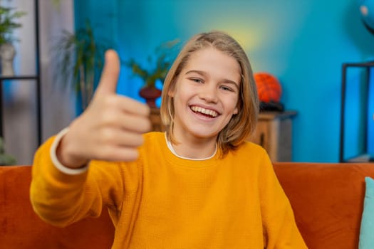 Like. Caucasian teenager boy gives thumbs up to camera while smiling at home. Sits comfortably on sofa, appearing satisfied with the good positive result, displaying feelings of approval, contentment