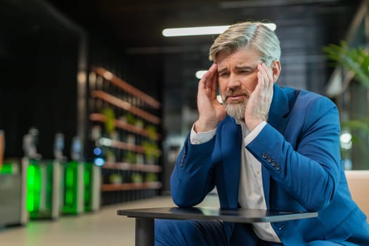 Tired ill middle-aged businessman suffering from headache problem tension and migraine, stress in modern office lobby. Handsome freelancer businessperson man guy sitting in hotel hall. Overworking.