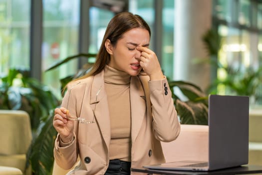 Overworked young Caucasian businesswoman suffering from eye strain and headache working on laptop sitting in office lobby. Tired female manager takes off glasses and massages, rubs bridge of nose.