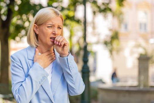 Sick mature businesswoman in formal suit suffering from sore throat while standing on city street. Upset Caucasian female manager with a cold or flu coughing and feels bad. She has a cold or virus.
