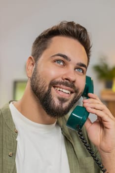 Closeup of smiling young man making wired telephone call conversation with friends sitting on couch at home. Happy excited guy male enjoying old-fashioned retro phone from 90s talking in living room.