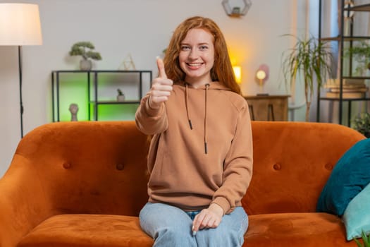Portrait of excited smiling Caucasian woman looking approvingly at camera showing double thumbs up, like sign, good news, positive feedback. Happy redhead girl sitting on sofa in living room at home.