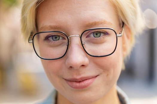 Close-up portrait of happy young woman face smiling friendly looking at camera resting relaxation feel satisfied good news outdoors. Pretty adult lady in glasses with short blond hair on city street.