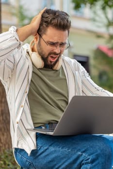 Sad young man tourist using laptop computer typing browsing, loses becoming surprised sudden lottery results, bad news, fortune loss, fail. Angry guy sitting in urban city outdoors. Town lifestyles.