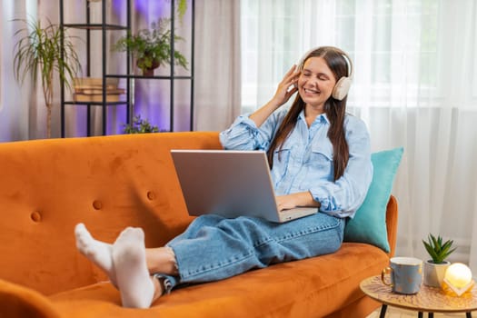 Happy relaxed Caucasian young woman in wireless headphones lying on home couch choosing listening disco music while working online on laptop netbook. Girl weekends activities entertaining. Lifestyles.