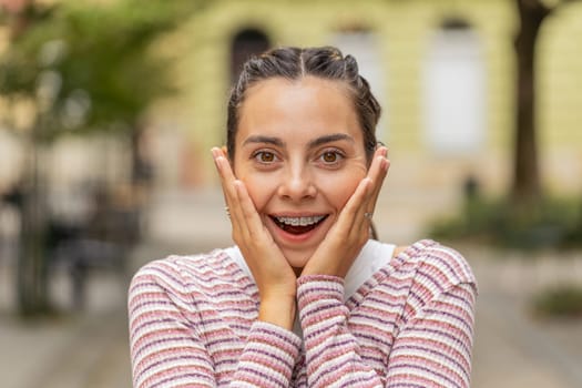 Portrait of excited Caucasian young woman tourist with braces on teeth walking on urban city street daytime outdoors. Girl expresses amazement, wow emotion, positive good mood enjoying sightseeing