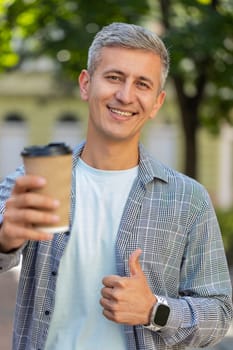 Happy Caucasian man enjoying morning coffee hot drink and smiling. Relaxing, taking a break. Mature guy standing on urban city center street, drinking coffee to go. Town lifestyles outside. Vertical