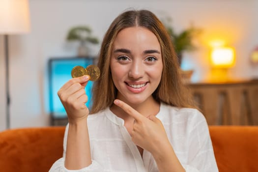 Portrait of happy woman holding two gold BTC coins at home successful developer programmer. Girl stock trader earning bitcoins after online monitoring trading operations. Increasing wealth. Lifestyle.