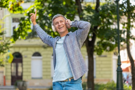 Happy relaxed overjoyed gray-haired mature man in wireless headphones listening disco music dancing outdoors. Cheerful adult guy in casual clothes walking in urban city street park. Town lifestyles.